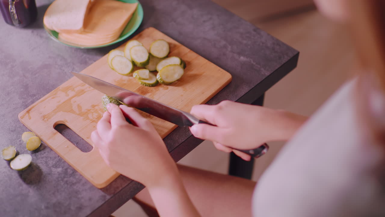 Overhead view of woman slicing cucumber on wooden cutting board in kitchen with bread and cheese on green plate beside coffee cup, preparing fresh meal with careful hands and sharp knife
