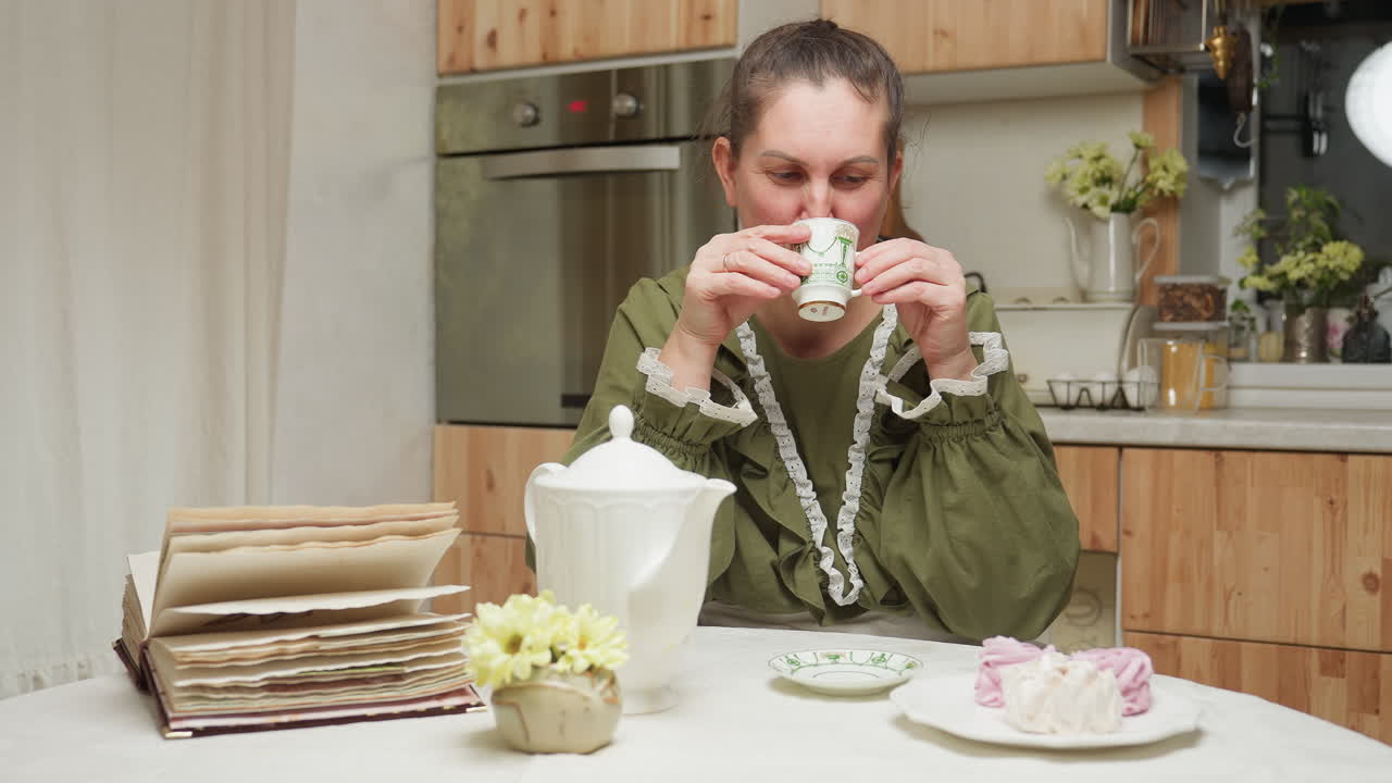 White lady drinks coffee from glass cup in cozy kitchen setting, then lowers cup slowly while gazing calmly at camera with relaxed expression, enjoying peaceful moment in morning