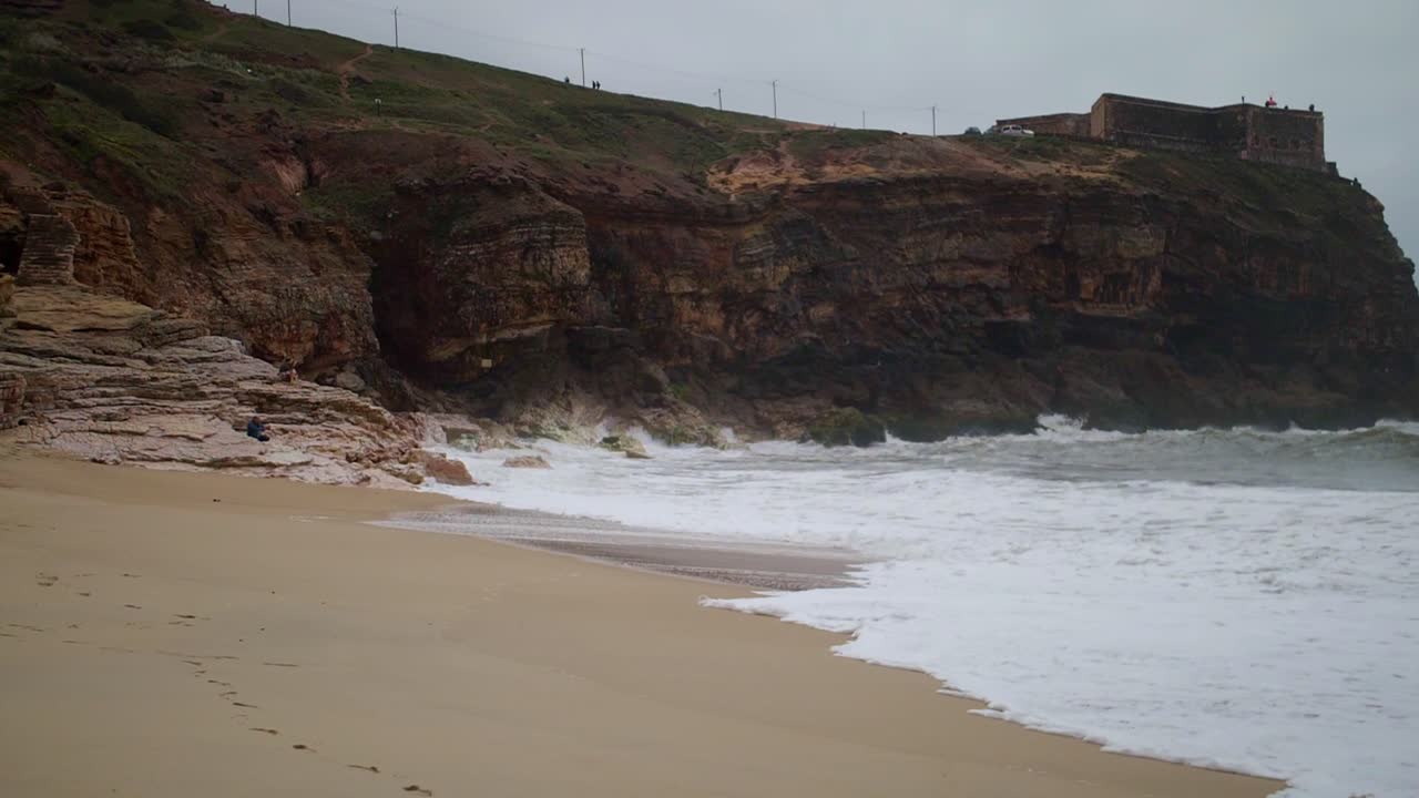 olas en cámara lenta blancas con espuma rompen en un acantilado rocoso de arena con un antiguo castillo en las montañas oscuras, cabo roca portugal