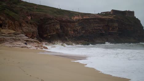 SLOW-MOTION-waves-white-with-foam-break-on-a-sandy-rocky-cliff-with-old-castle-on-the-dark-mountains,-Cape-Roca-Portugal