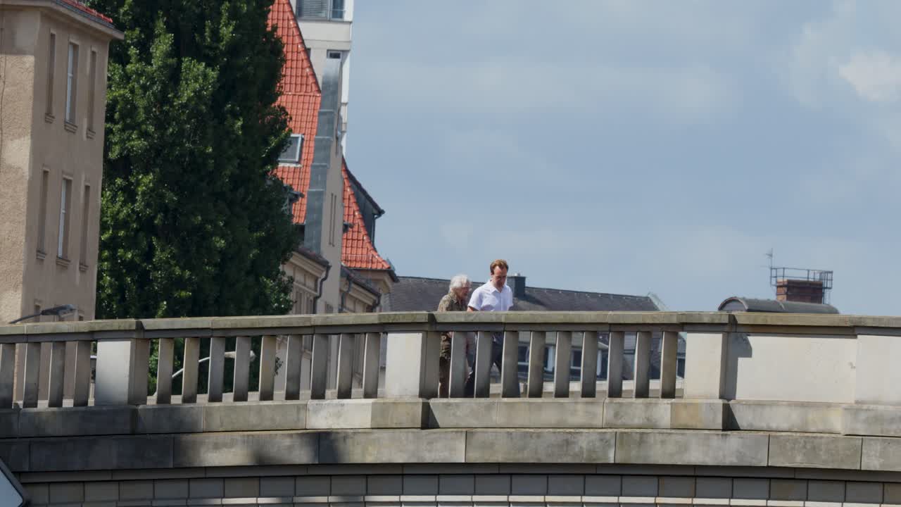 Two adults walk and talk on a stone bridge, sunny daylight, static camera, urban backdrop