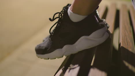 close-up of single foot wearing black canvas sneakers with white soles resting on a wooden bench outdoors at night under soft lighting, with blurred background