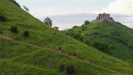 two people running uphill on a green trail with an ancient fort in the background
