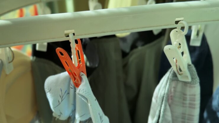 Detailed close-up of clothespins gripping small garments on an indoor laundry rack, lit by soft daylight near a window, showing a quiet household drying routine inside an apartment