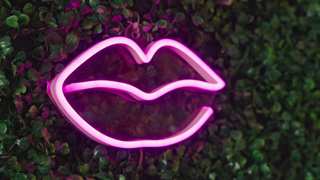 Insegna a forma di labbra al neon rosa che si illumina su una parete di fogliame verde.