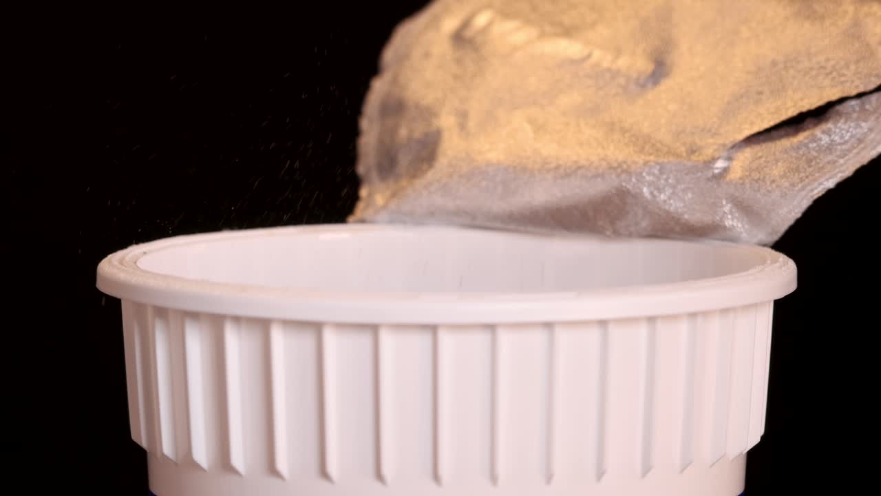 A hand pours spice powder from a packet into a white instant noodle cup against a black background, with even studio lighting and static camera