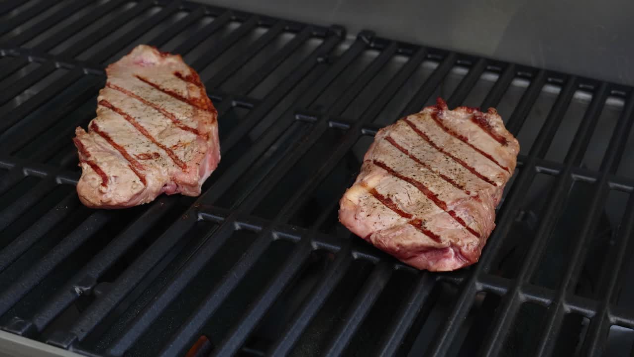 Two beef steaks sizzle on a grill, showcasing perfect grill marks and even cooking in a well-lit kitchen setting