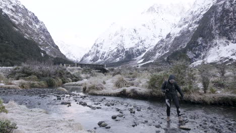 foto em câmera lenta de garota em equipamento de caminhada cruzando o rio cercado por montanhas cobertas de neve