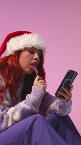 Vertical-Video-Studio-Shot-Of-Young-Gen-Z-Woman-Wearing-Christmas-Santa-Hat-Eating-Candy-Lollipop-Looking-At-Mobile-Phone-3