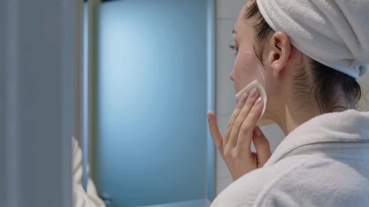 Bringing cotton pad, woman steadying jaw cleansing cheek at vanity, wearing white robe, copy space