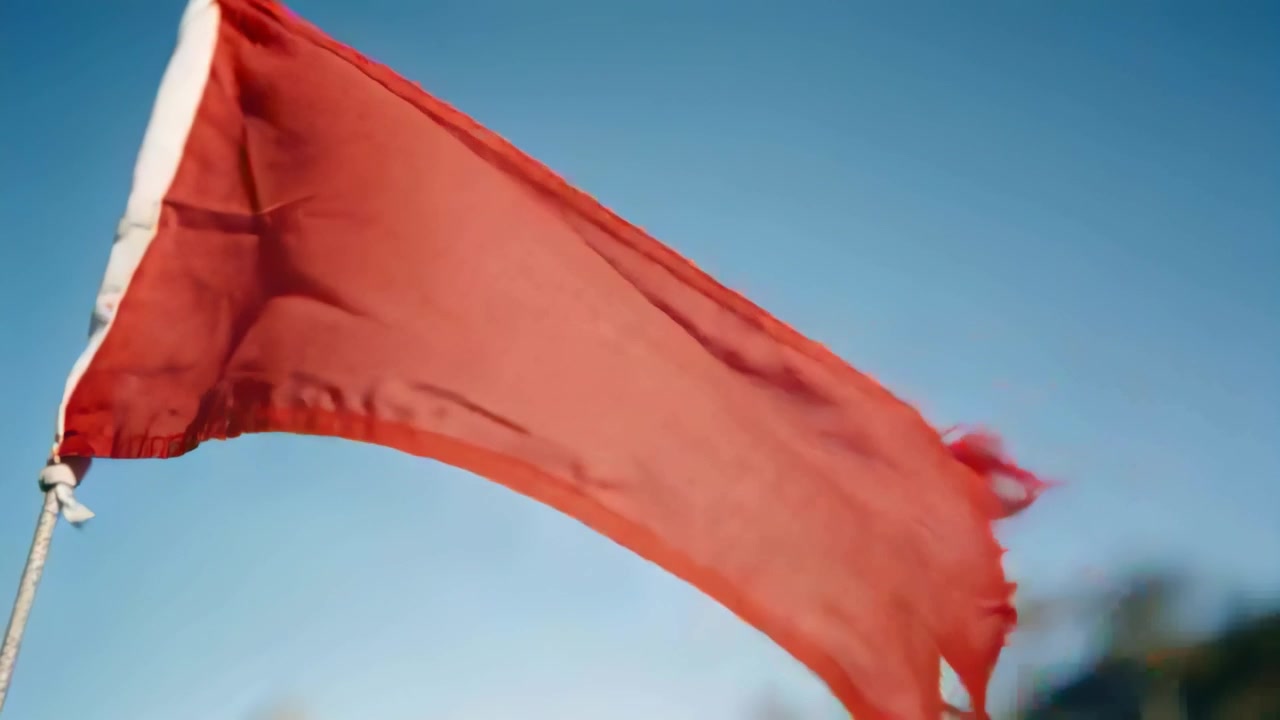 Viento racheado levantando y luego amainando una bandera roja ondeando con el borde deshilachado cerca de los nudos de izado sobre un campo abierto.
