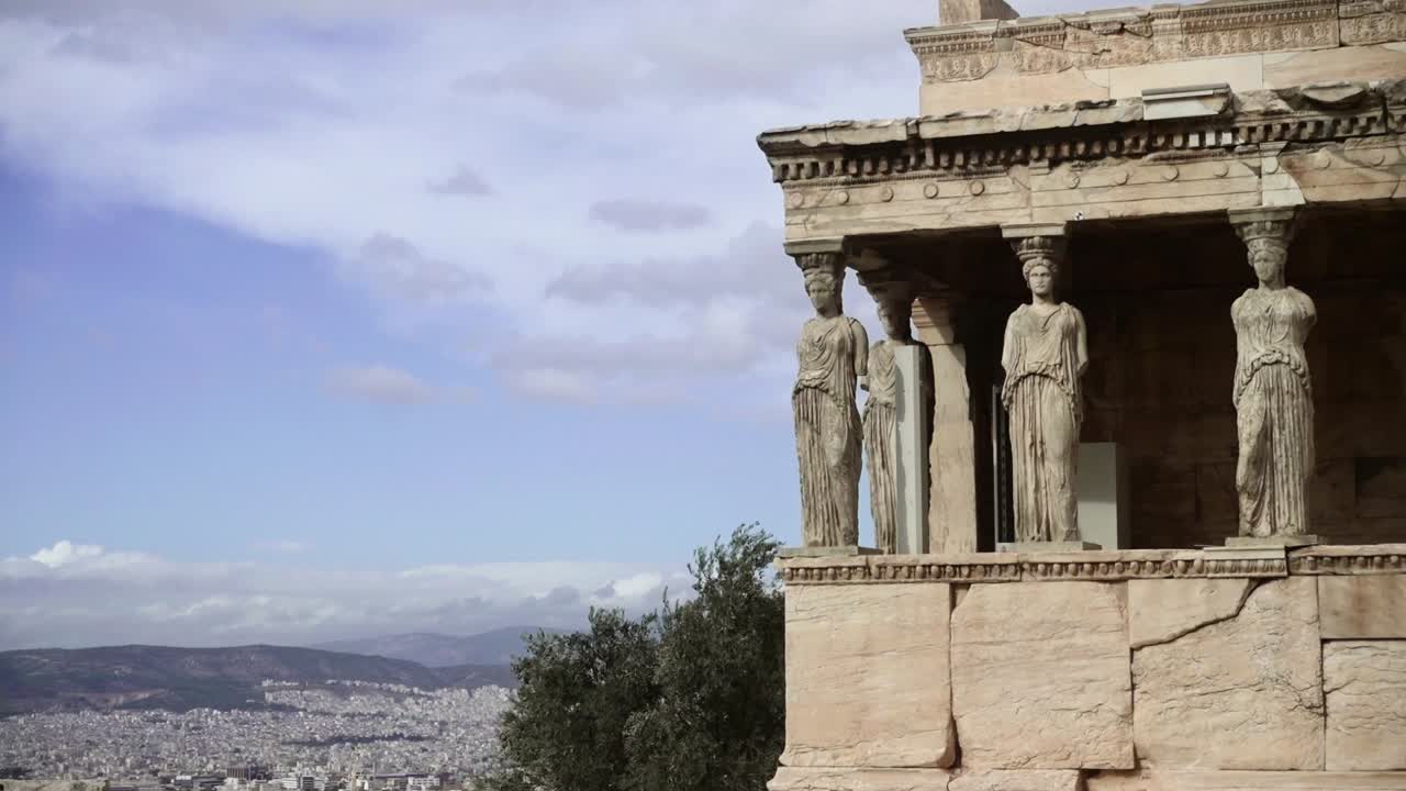 Pan down along the stone walls outside of the Porch of the Caryatids by the Erechtheion down to the city of Athens on a hot day in Greece