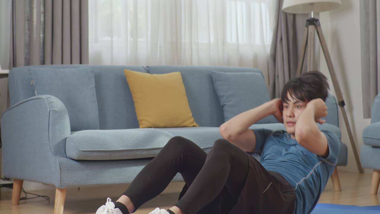 스포츠 옷을 입은 운동 체력 있는 아시아인 남자가 넓고 밝은 거실에서 가정에서 복부 걱거리는 운동을 하고 있습니다.