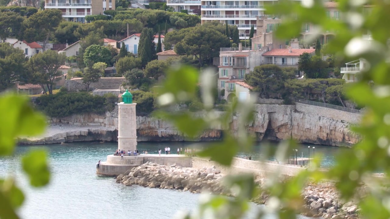 Inquadratura statica del faro di Cassis e degli edifici costieri, incorniciati da foglie verdi sfocate, Francia.