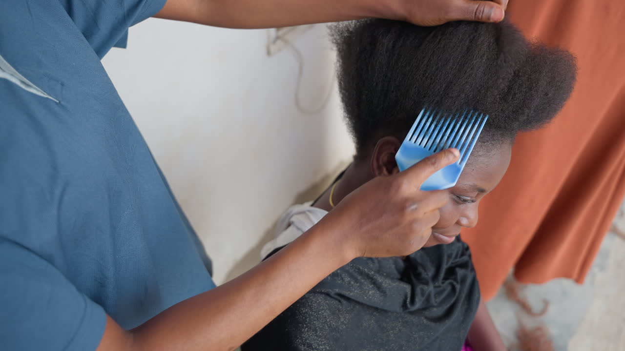 Sort kvinde får omhyggelig hårstyling i hjemmesalon, stylist og lærling reder og udreder forsigtigt afrohår, hænder deler håret op og inspicerer hovedbundens tekstur, nærbillede af krøller mod et varmt gardin.
