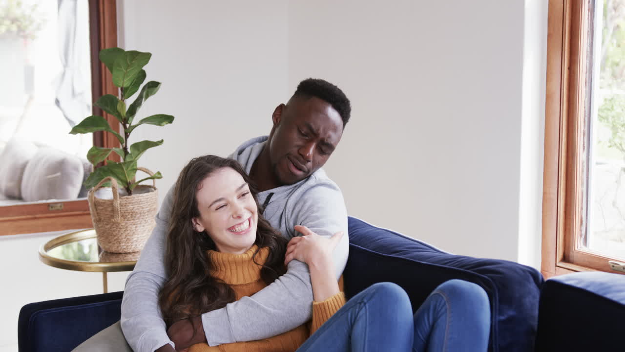 Happy diverse couple sitting on sofa laughing and embracing in home,copy space