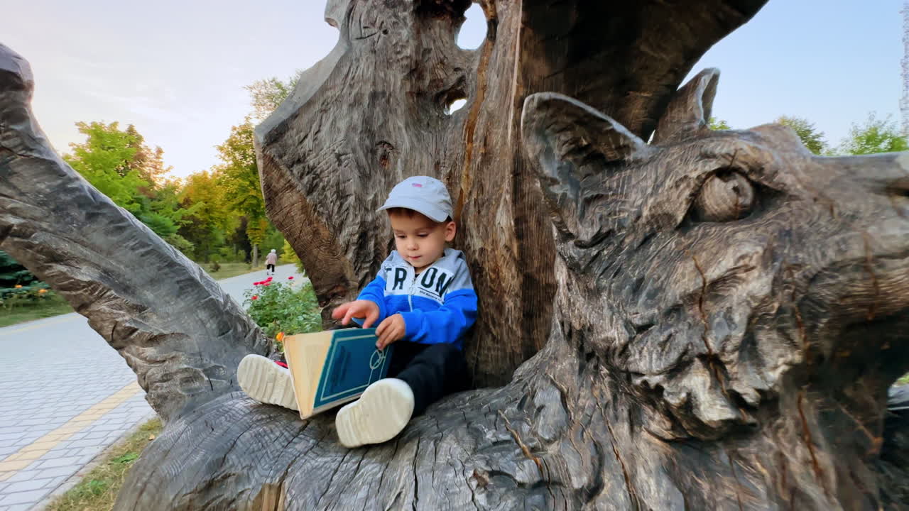 Little boy sitting on the cat statue carved from wood. Cute toddler is focused on the book.