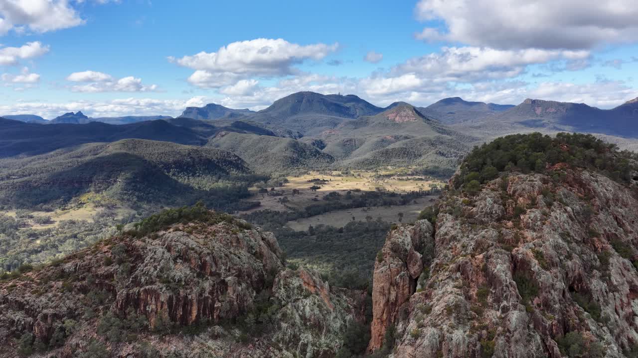 Drone camera smoothly pans over rugged Split Rock, revealing expansive mountain ranges and valleys under bright daylight in Warrumbungle National Park, Australia