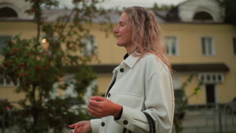 cheerful student walking along urban sidewalk twirling closed umbrella with soft smile, wearing light jacket, relaxed expression, residential buildings and parked vehicles in background