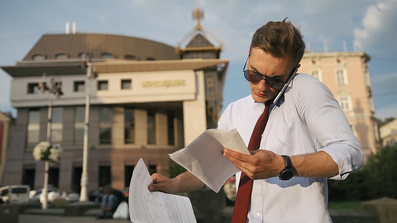 Man talking on phone while holding documents in front of a building