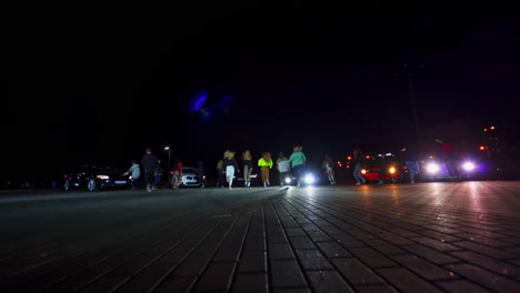 group of young dancers moving outdoors at night time. teenagers run to the race cars standing at backdrop. low angle view.