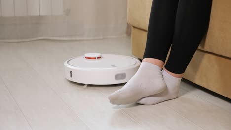 woman in white socks lets robot cleaner pass sitting on sofa