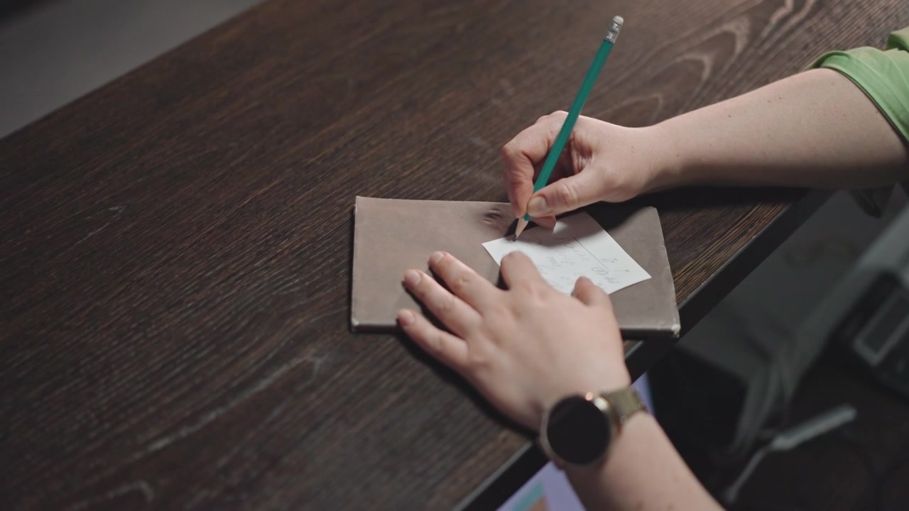 Mãos vistas de cima escrevendo em caderno em mesa de madeira escura, smartwatch no pulso, professor preparando aula e corrigindo diagramas de acordes, papelaria organizada e envelope, planejamento focado para a prática do aluno