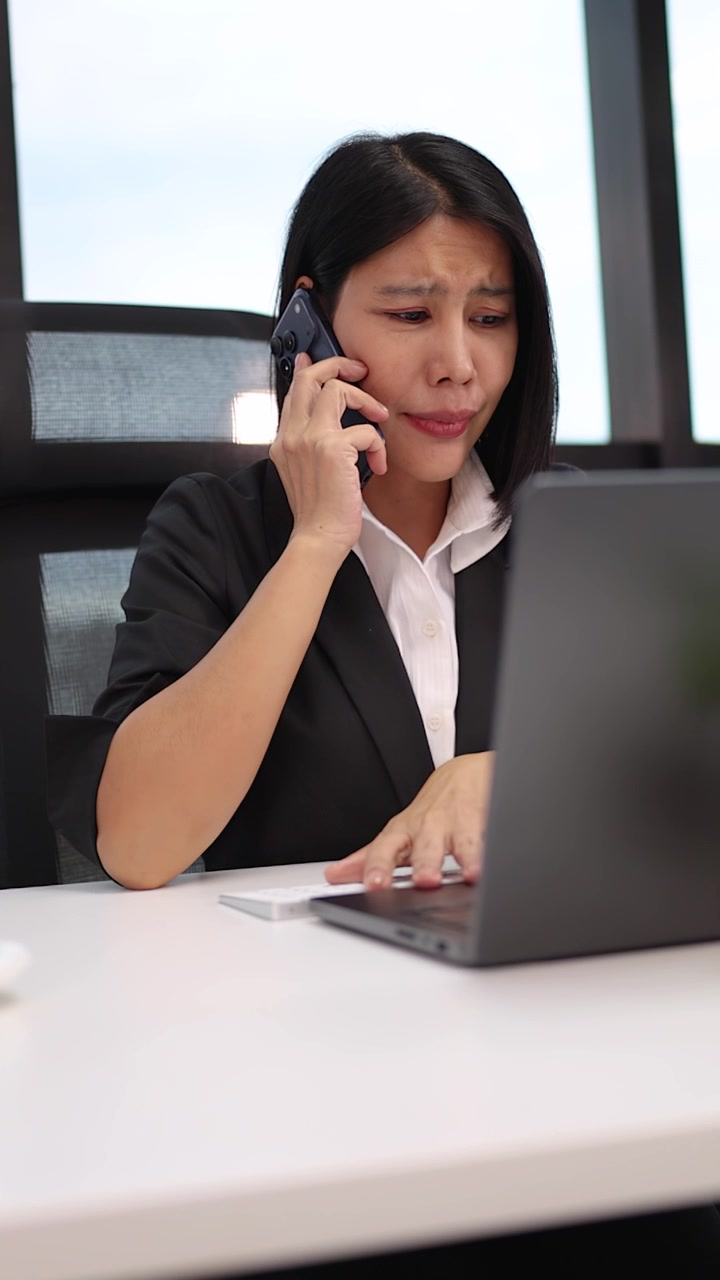 Znepokojená podnikatelka emocionálně reaguje na stresující telefonát, zatímco pracuje na notebooku ve světlé kanceláři