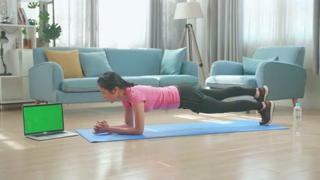 mujer asiática haciendo plancha de yoga y viendo tutoriales en línea en una computadora portátil de pantalla verde, entrenando en la sala de estar