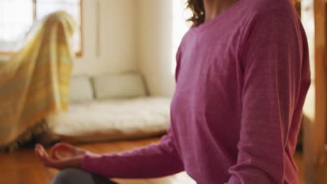 Relaxed-mixed-race-woman-practicing-yoga,-sitting-in-meditation-in-sunny-cottage-bedroom