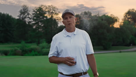 rich golf player stand relaxed on course. old man smoking cigar holding drink.