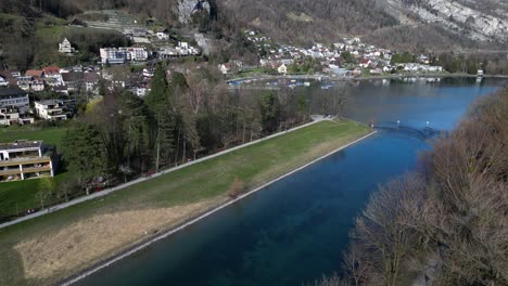 Drone-clip-over-calm-straight-river-at-the-foothills-of-the-Swiss-Alps,-running-past-a-traditional-village