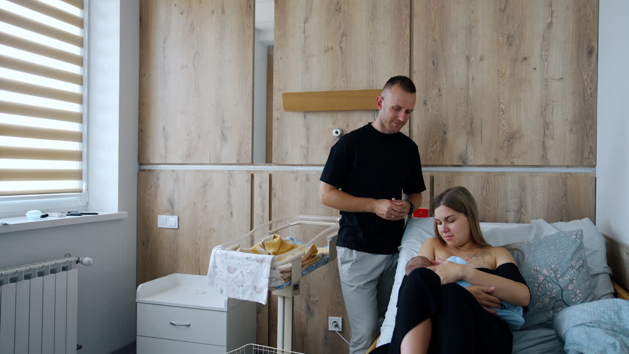 New-made parents in the after-labor ward in maternity hospital. Mother is breastfeeding her newborn and dad stands watching beside.