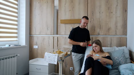 new-made parents in the after-labor ward in maternity hospital. mother is breastfeeding her newborn and dad stands watching beside.