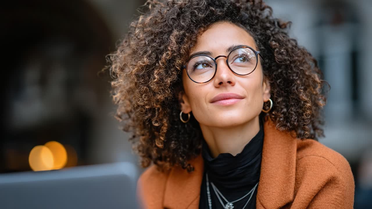 Thoughtful Young Woman with Curly Hair and Glasses Expresses Curiosity While Working on a Laptop in a Cozy Setting