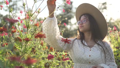 retrato de una niña con gafas, sombrero, vestido blanco, sonriendo muy feliz y oliendo una flor en un campo de flores de zinnia, durante la puesta de sol