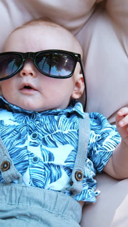 funny baby boy wearing mom's sunglasses. kid wearing a blue shirt and shorts lies on a soft pillow outdoors.. vertical video