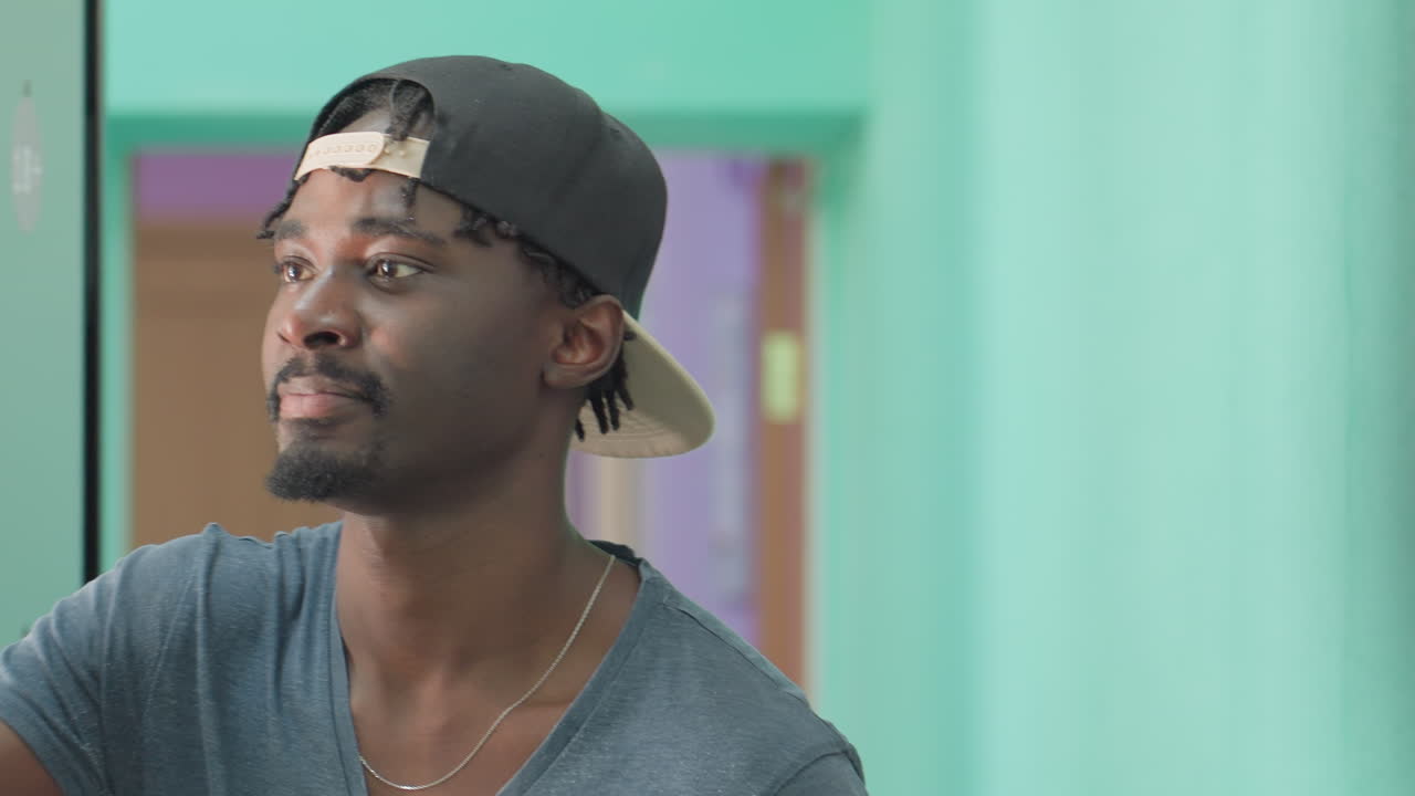 Close up of happy young man in casual cap dancing joyfully while reaching toward shelf to take book in bright indoor hallway with colorful walls, showing lively mood