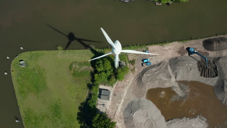 Una-Estructura-De-Motor-Eólico-Girando-Sus-Palas-Cerca-Del-Canal-Gouwe-Con-Vista-A-Una-Excavadora-Hidráulica-En-Un-Sitio-De-Construcción.