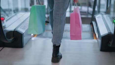 back view of woman in blue jeans and black boots stepping onto descending escalator, holding two shopping bags, blurred background in modern mall setting