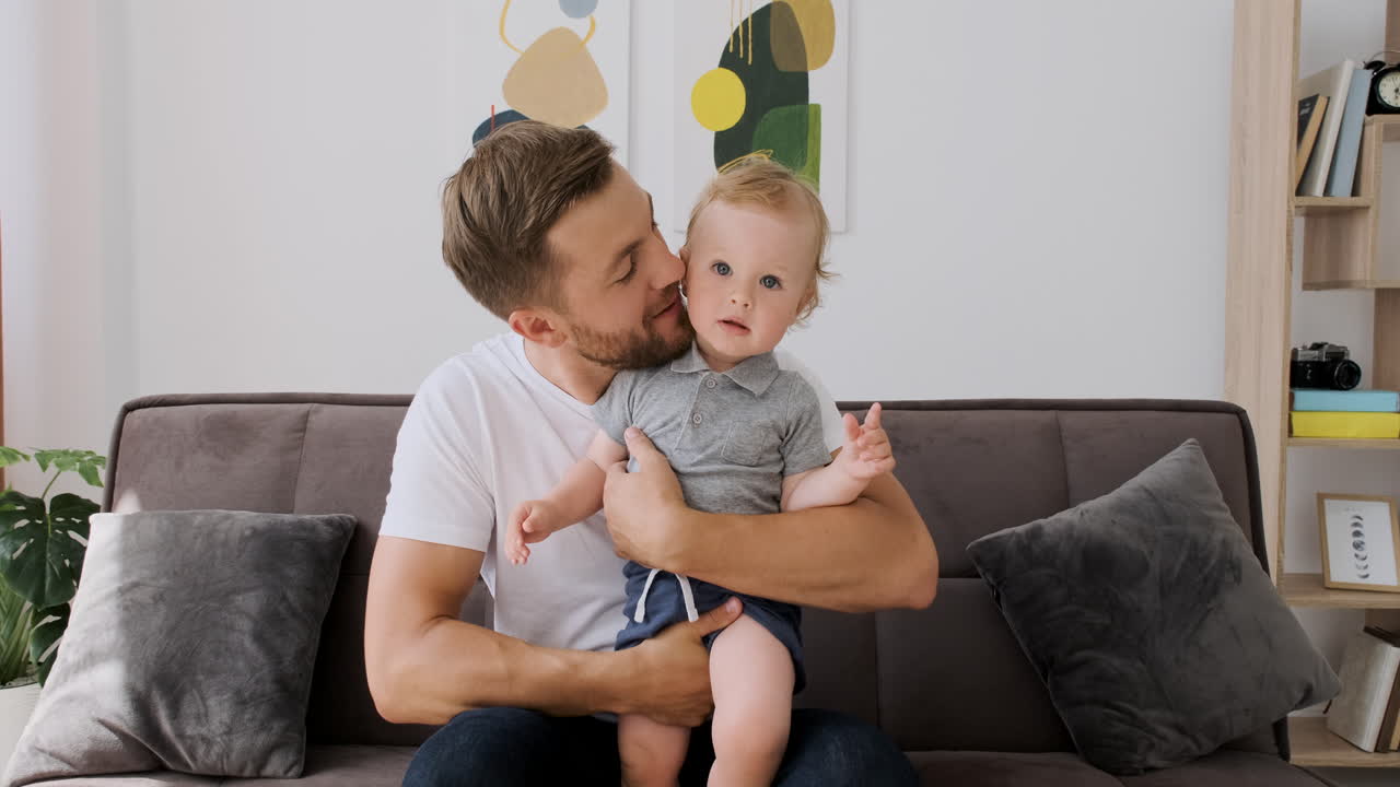 Handsome Father Sitting On Sofa In Living Room Holding And Kissing His Cute Child While Talking On Video Call