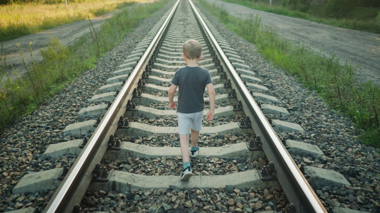 back view of boy in casual outfit walking alone on rail track surrounded by gravel and vegetation, long perspective view stretching into distance, calm rural setting with roadside