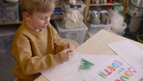 niño rubio dibujando con lápiz verde sentado en una mesa en un taller de artesanía donde hay carteles con citas ambientales