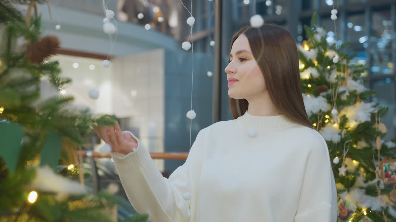 Woman in white sweater admires Christmas tree decorations, gently touching ornaments, surrounded by twinkling lights and festive decor in a mall