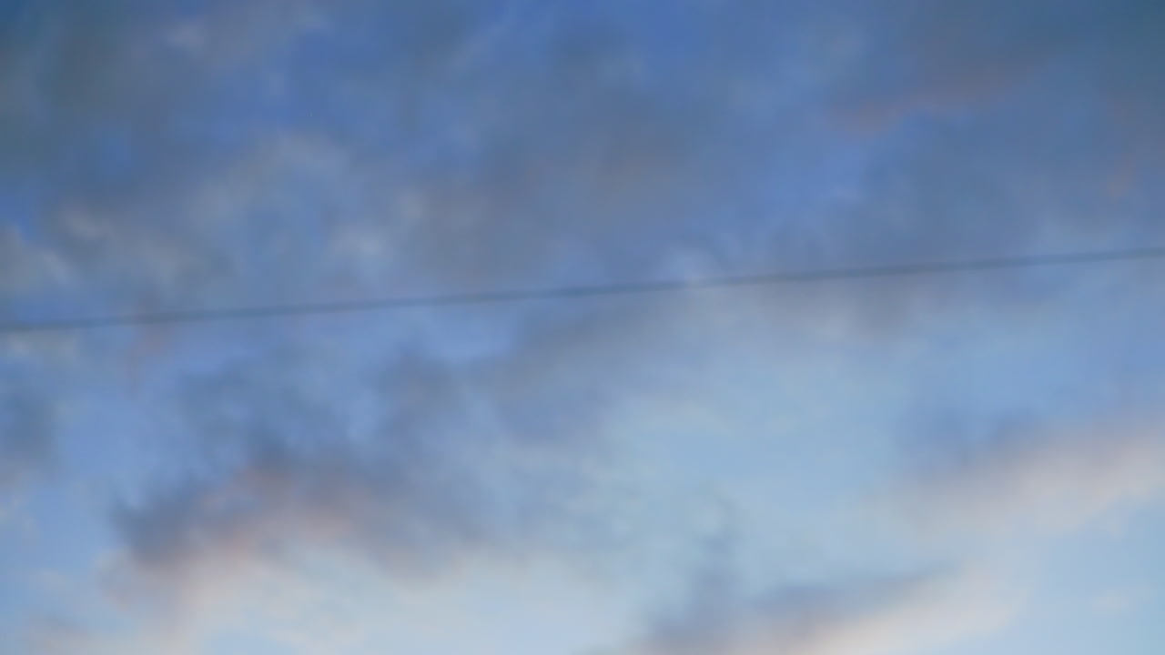Evening sky view with soft drifting clouds over urban setting featuring moving cars, parked vehicles, and faint outline of office buildings, capturing calm atmosphere of city life at dusk