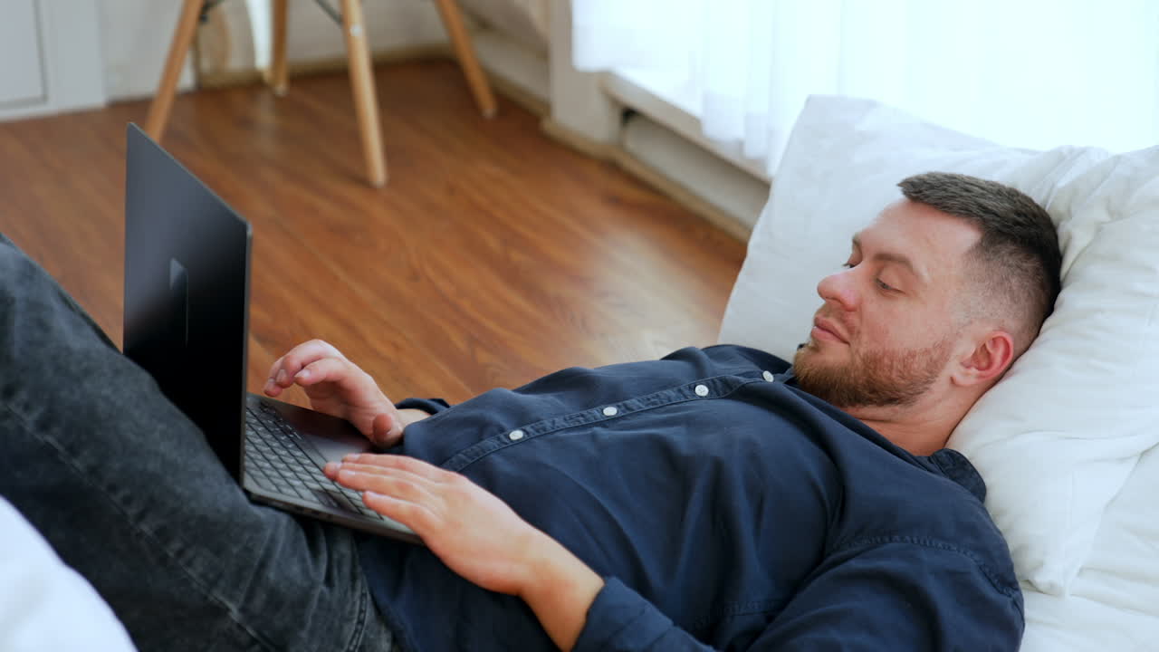 Peaceful relaxed man lying on the pillows looks at his laptop. Man using gadget for entertainment.