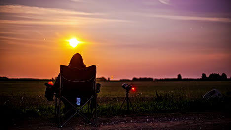 person in a hoodie along a countryside road watching the new moon rising in a camping chair capturing a time lapse