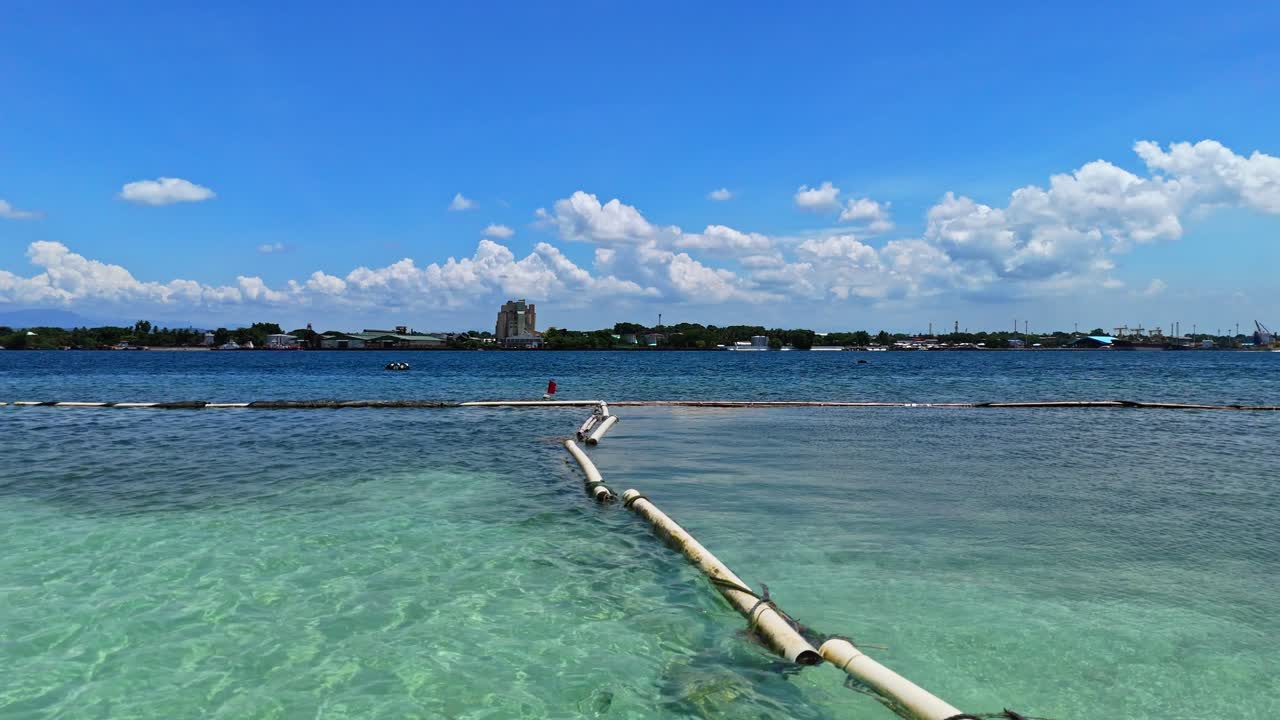 Clear drone view of shallow turquoise waters, floating barriers, and distant coastal buildings under bright skies—perfect for travel, maritime, and documentary use