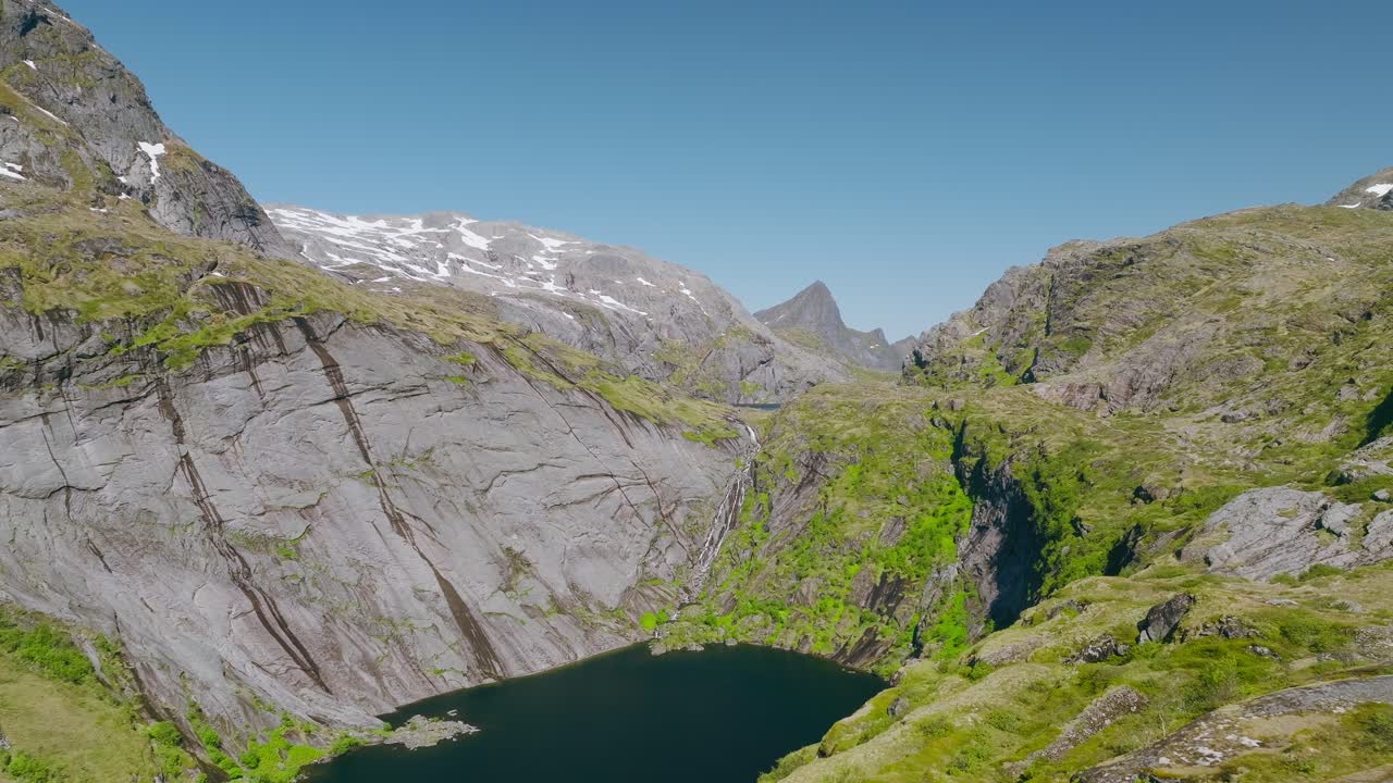 Un uomo e una donna si trovano sul bordo di una scogliera di montagna che si affaccia su un lago sottostante, circondati da montagne di granito innevate, catturando un momento alpino drammatico e sereno nelle Lofoten, Norvegia, durante l'escursione a Munken.