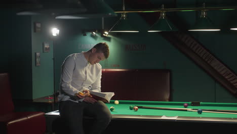 concentrated man in white shirt and plaid pants sits on green pool table, reading book while rolling billiard ball in hand. pool cues and scattered billiard balls add to the relaxed ambiance